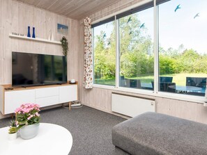 A modern living room with a flat-screen TV, a grey sofa, a white coffee table, and a large window with a view of greenery and birds. - 5 Person Holiday Home in Bording (Bording)