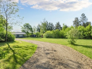 En grusvej fører til et hus med metal tag, omgivet af frodig vegetation og træer.