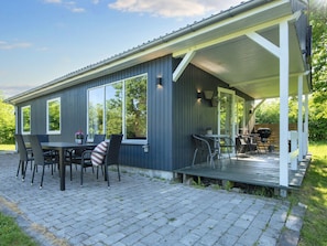 Een overdekt terras buiten met een tafel, stoelen, een barbecue en uitzicht op het omliggende groen.