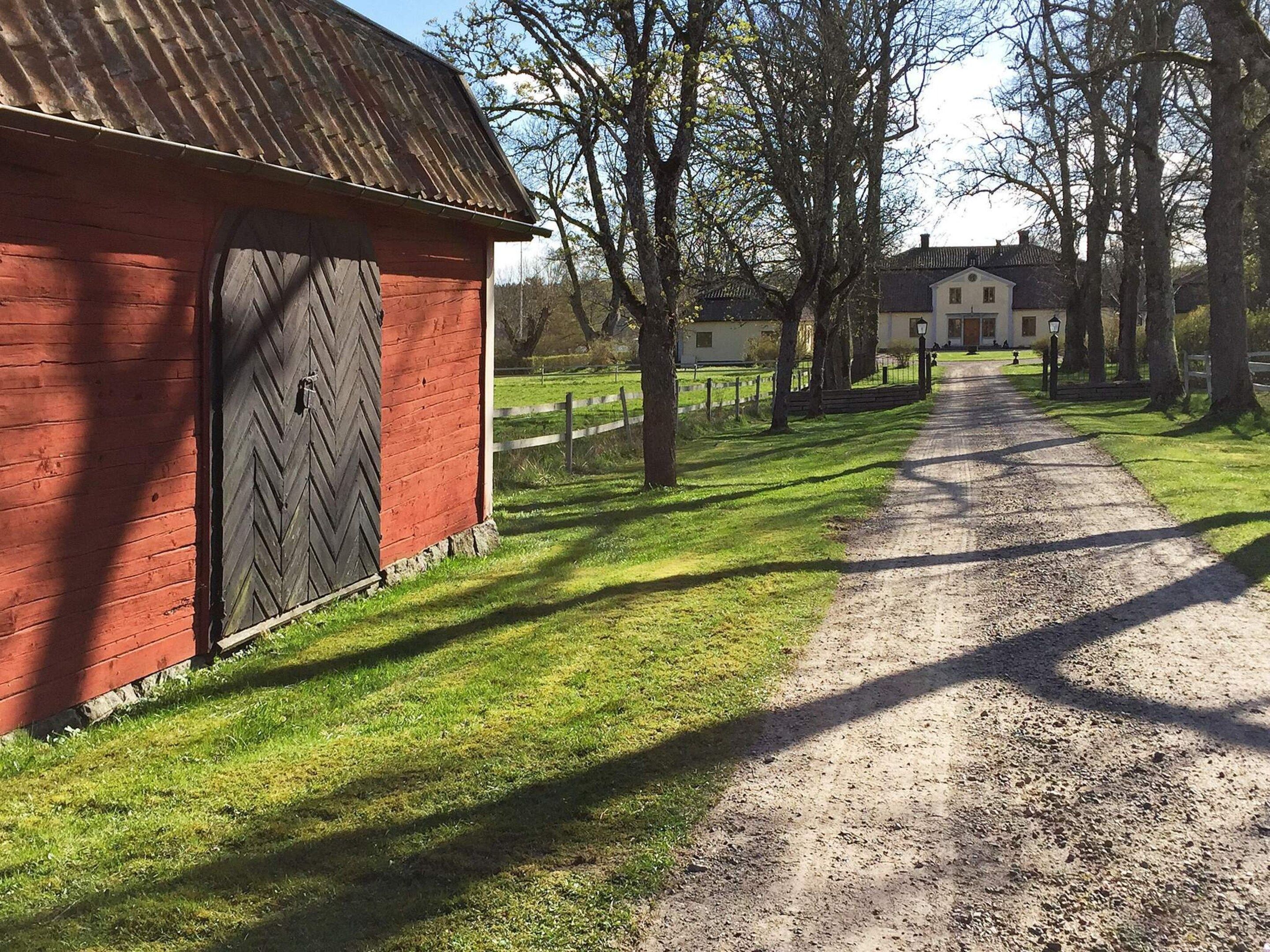 A rural road leading to a red barn with a large door, surrounded by trees and a grassy area.