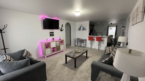 A modern living room with grey sofas, a coffee table, and a TV. There's a kitchen area with a bar and red stools. - Welcome to Innovators Home Broken Arrow with Large Driveway (Broken Arrow)
