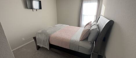 Un dormitorio con cama, televisión montada en la pared y ventana con cortinas.