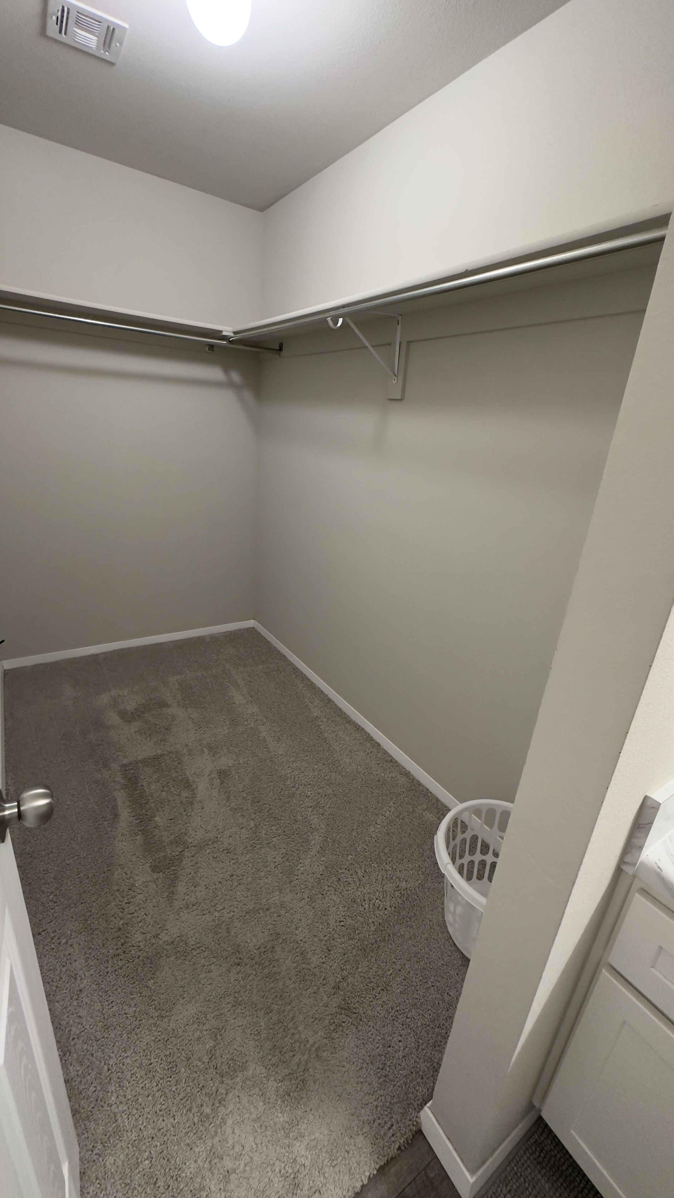 Una habitación pequeña con alfombra, una puerta blanca, un respiradero en el techo y un cesto de ropa blanca.
