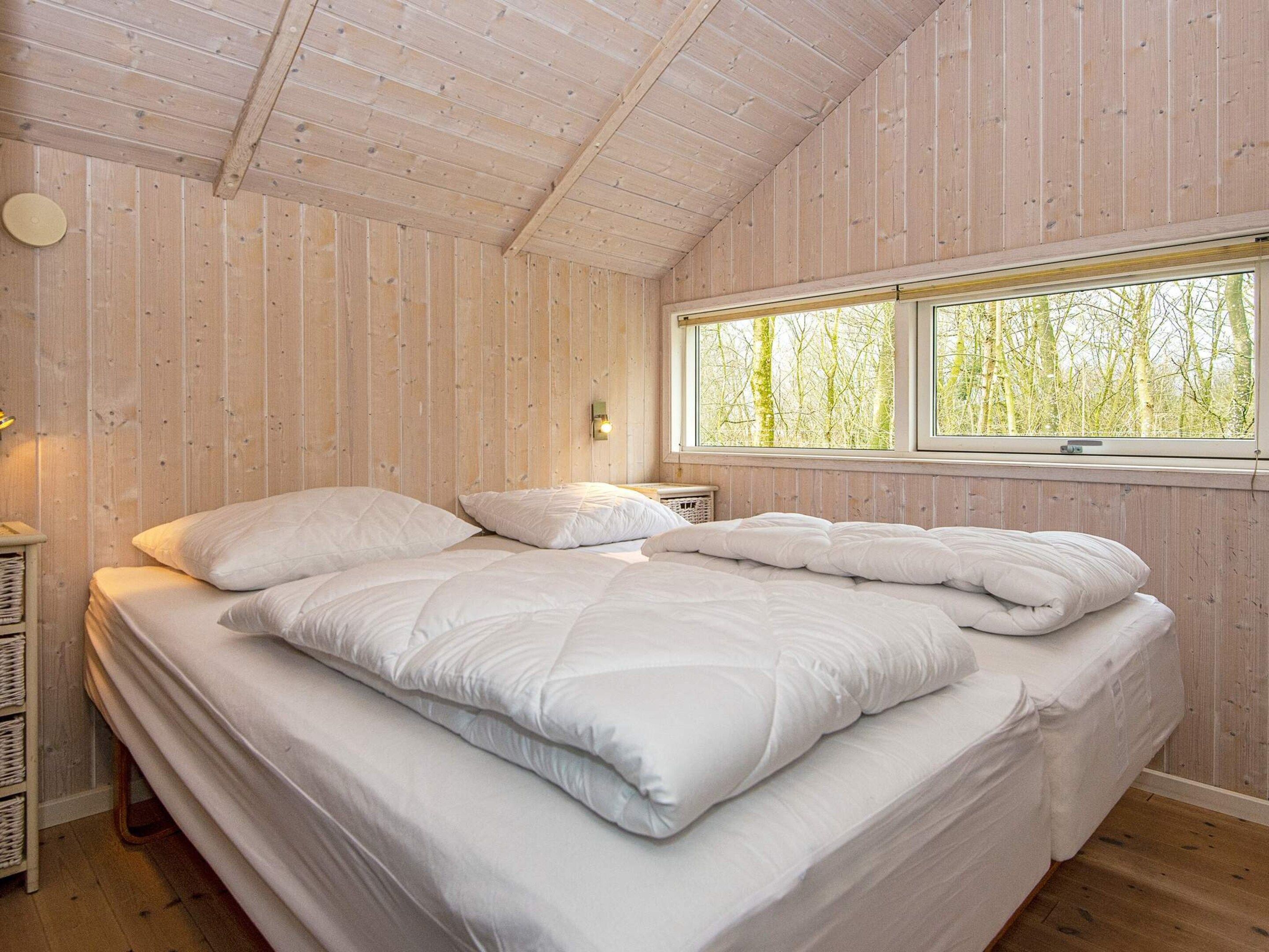 A bedroom with a wooden ceiling, a large bed with white bedding, a window with a view of trees, and a wall-mounted light fixture.
