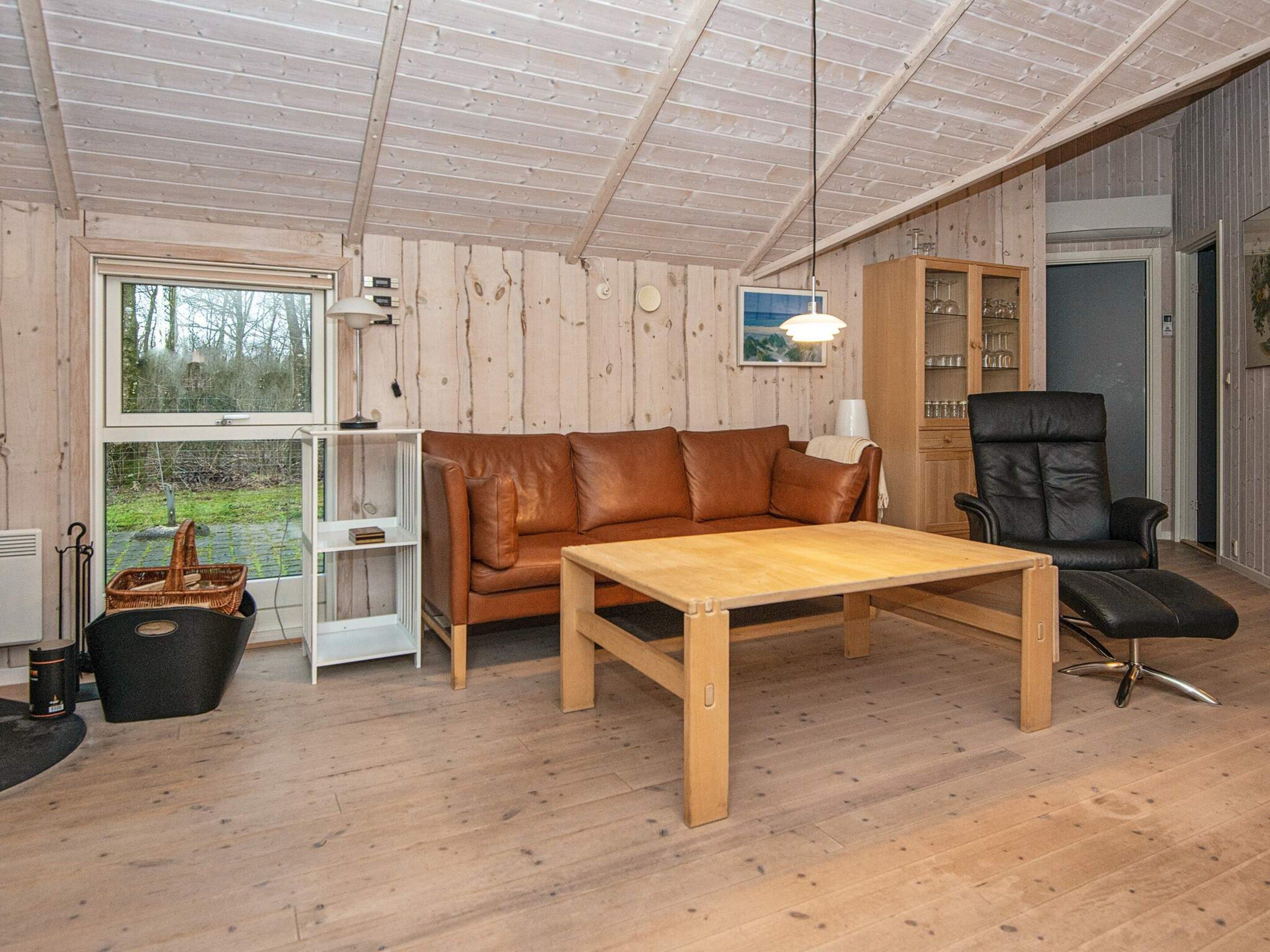 A cozy room with a wooden floor, a brown leather sofa, a wooden table, a black leather chair, and a window with a view of greenery.