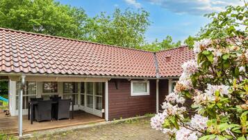 A house with a tiled roof, a covered patio area with outdoor furniture, and a garden with flowering shrubs.