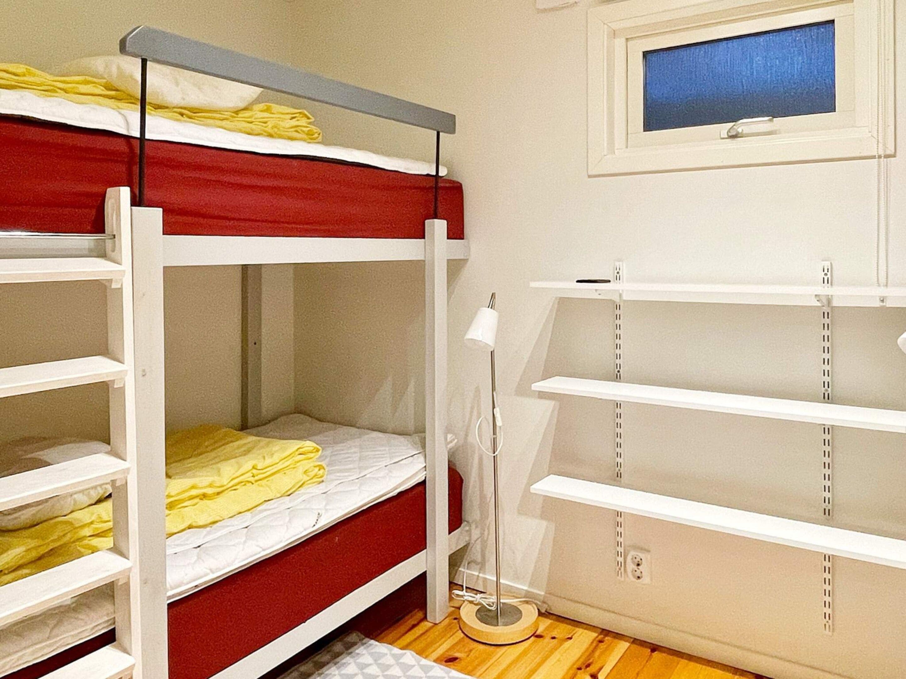 A bunk bed with a red mattress and white bedding, a bedside table with a lamp, and a window with a blue view.