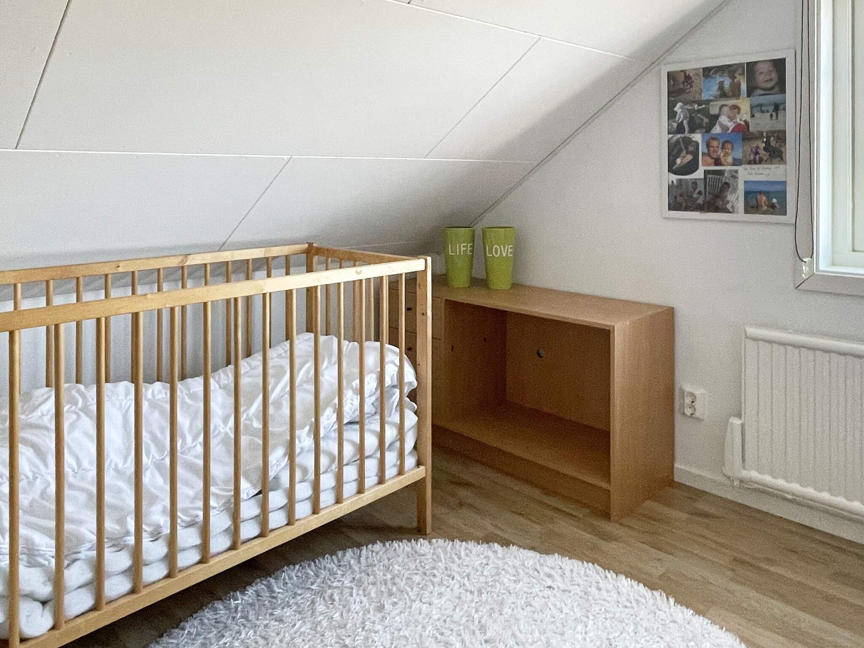 Une pièce avec un berceau en bois, un tapis blanc et moelleux, une étagère en bois avec des récipients verts et un mur orné d’un collage de photos.