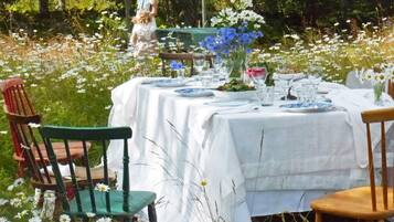 Un espacio exterior con comedor, mesa lista para dos personas, sillas circundantes, jardín con flores y árboles.
