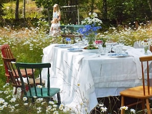 An outdoor dining area with a table set for two, surrounded by chairs, and a garden setting with flowers and trees. - 6 Person Holiday Home in Lidkoping (Lidköping)