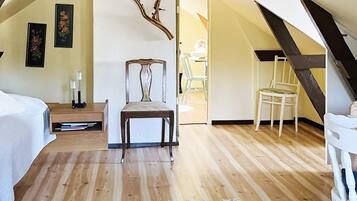 Una habitación con techo inclinado, suelo de madera, cama, silla, una mesita y cuadros colgados en la pared.