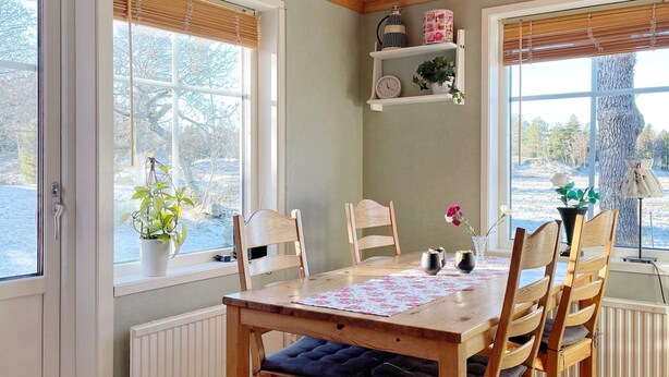 A dining area with a wooden table and chairs, a window with a view of trees, and a shelf with decorative items. - 6 Person Holiday Home in Lidkoping (Lidköping)