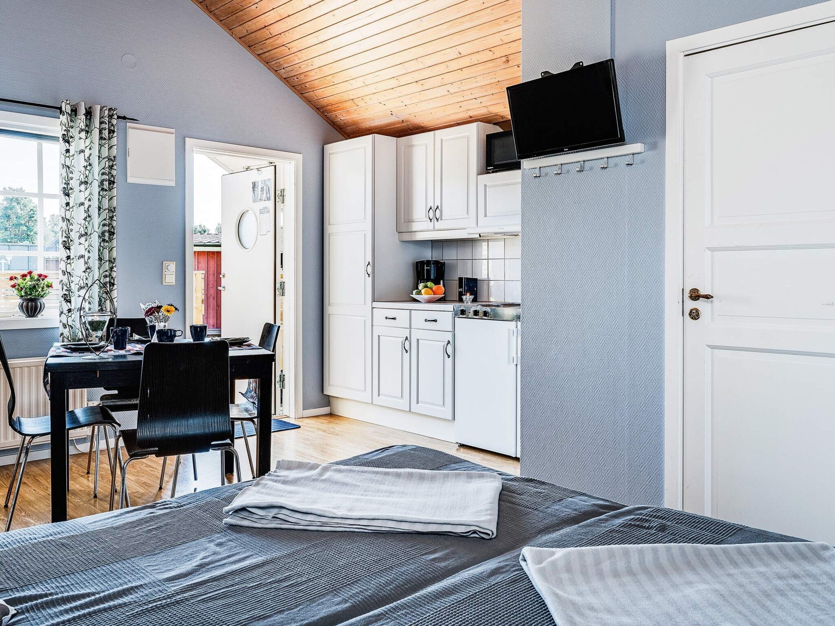 A compact room with a bed, a dining table with chairs, a kitchenette, and a television mounted on the wall.