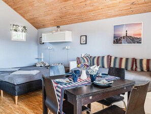 Een gezellige woonkamer met een houten plafond, een zithoek, een eettafel met stoelen en een groot raam.