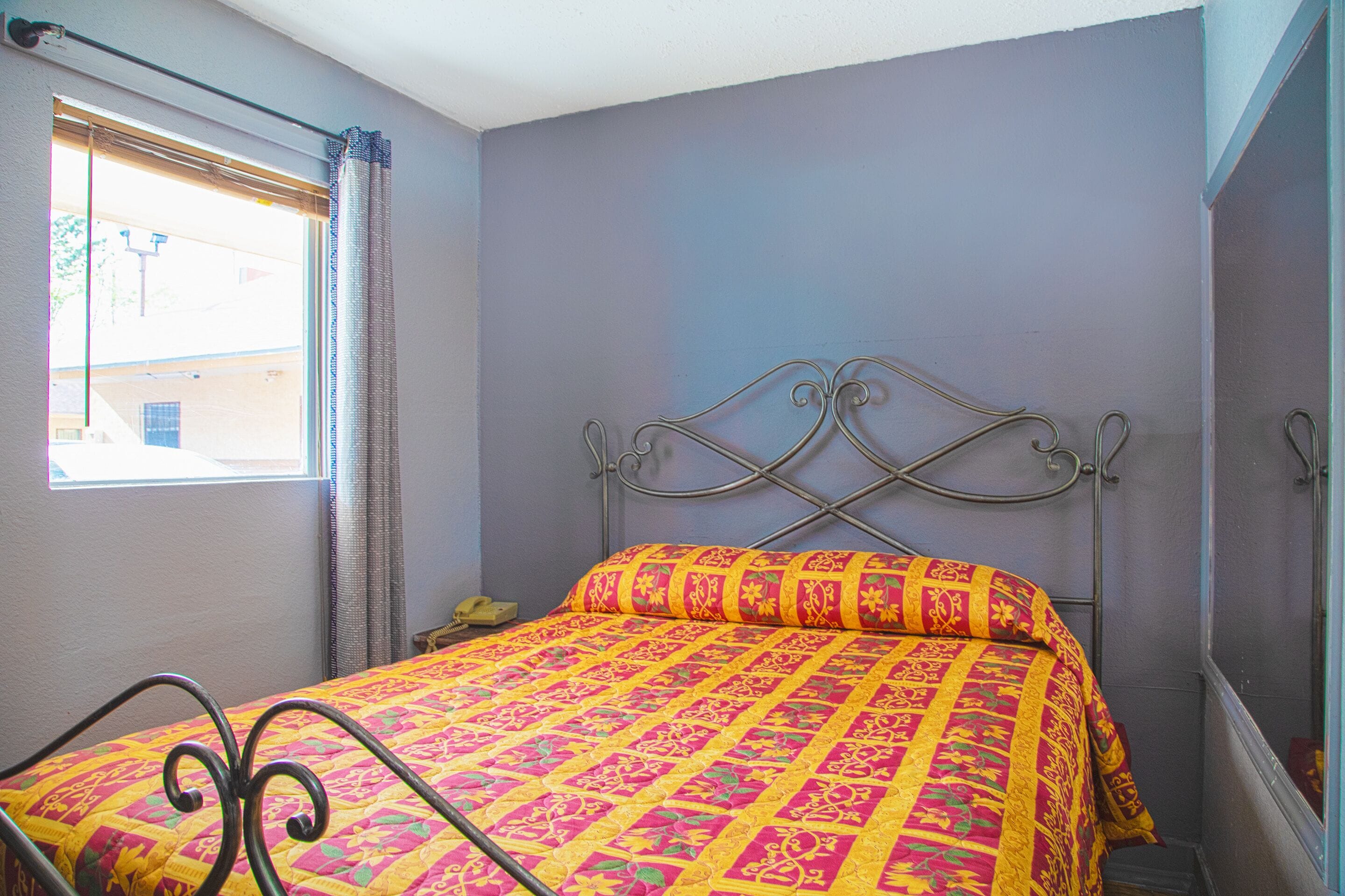 A bed with a metal frame and a colorful quilt, a window with curtains, and a plain wall.