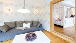 A modern living room with a grey sofa, white cushions, and a white coffee table with a decorative item. There are two pendant lights and a wall-mounted light fixture. A large mirror reflects a bedroom with a bed and curtains. - Ackerbürgerhof Ballstraße 18 (Quedlinburg)
