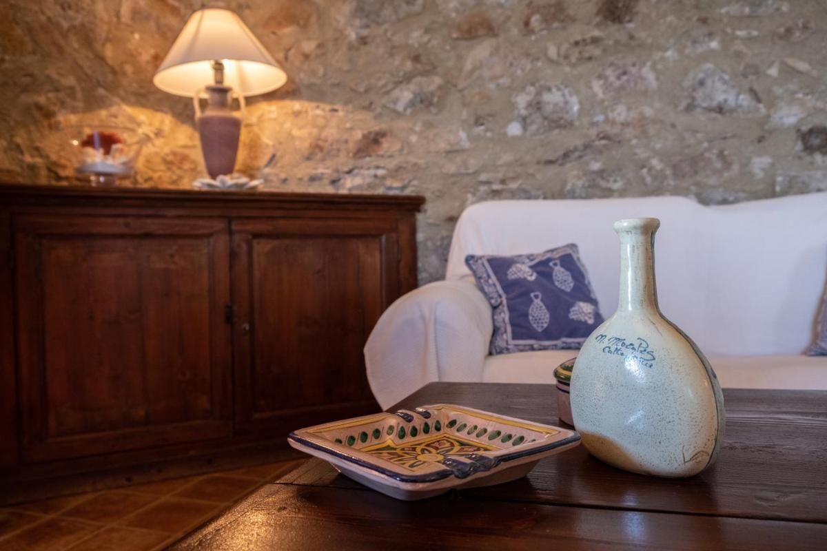 Une pièce chaleureuse avec un mur en pierre, une armoire en bois, un canapé blanc agrémenté de coussins à motifs, une table avec une assiette en céramique et un vase, et une lampe de table.