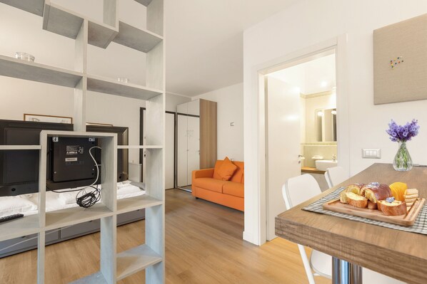 A modern living room with a sofa, a dining table with bread and fruit, and a TV on a shelf.