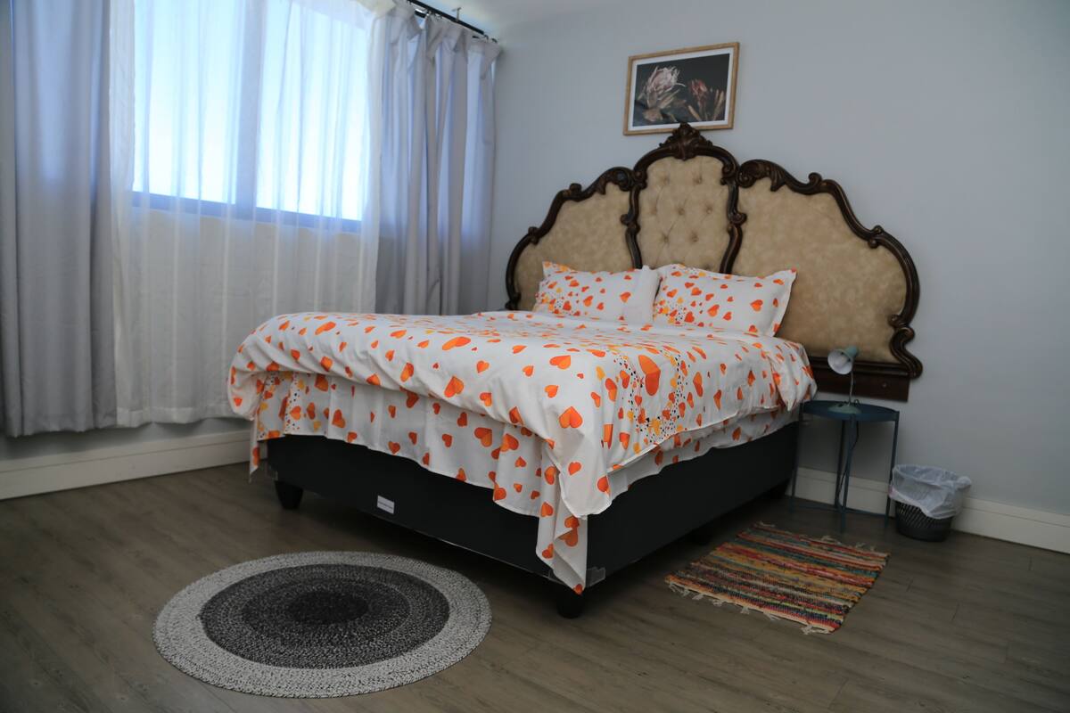 Quarto com cama, mesa de cabeceira, lixeira, janela com cortinas e um quadro na parede.