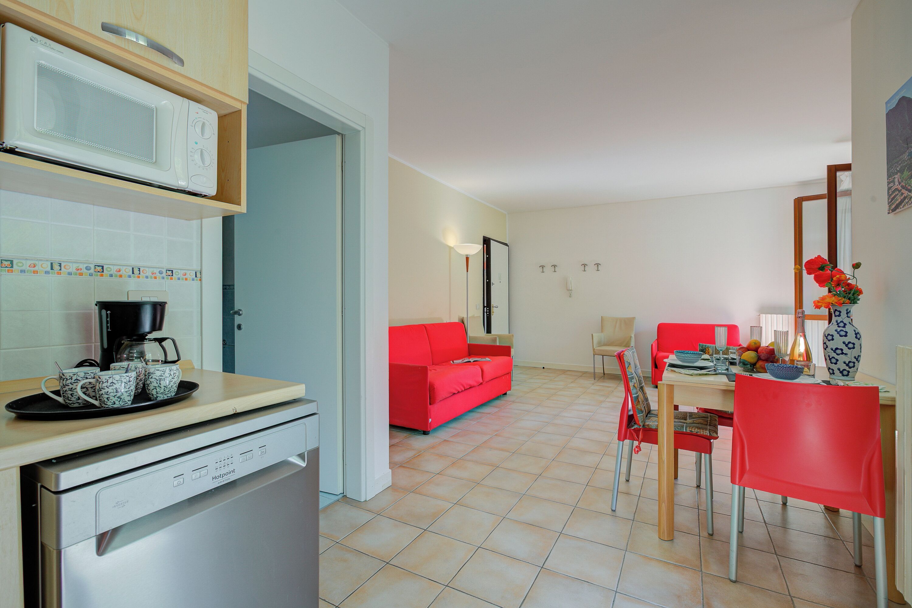 A kitchen with a microwave, coffee maker, and dishwasher; a dining area with a red sofa, chairs, and a table; and a living area with a red couch and a painting on the wall.