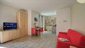 A living room with a red sofa, a TV on a wooden cabinet, a dining area with red chairs, and a kitchen in the background. - Porto Letizia Napoli 6, Porlezza, Italy (Porlezza)