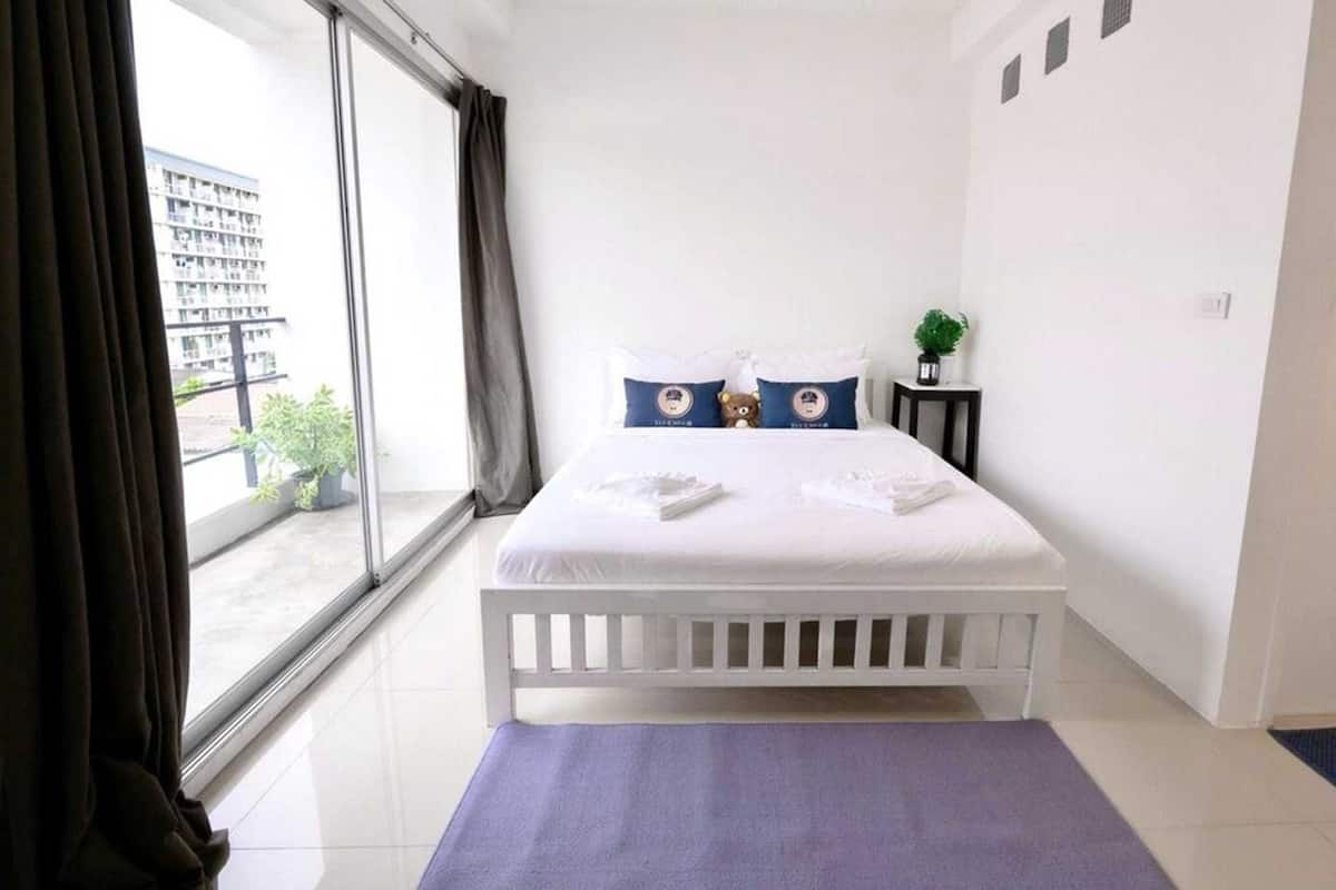 A hotel room with a large window, a white bed, a purple rug, and a small plant on a side table.