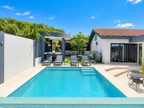 Une piscine rectangulaire, agrémentée de chaises longues et d’une table, entourée d’une terrasse avec un bâtiment et un espace salon couvert.