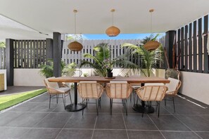 An outdoor patio with a wooden table and wicker chairs, surrounded by potted plants and a decorative fence.