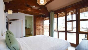 A bedroom with a large bed, wooden beams, a window, and a door leading to another room.