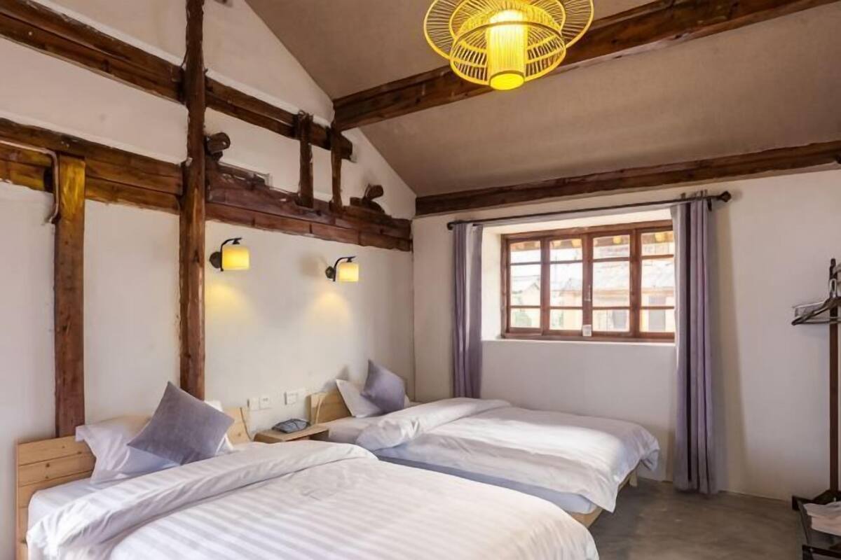 A room with two beds, wooden beams, a window with curtains, and wall-mounted lights.