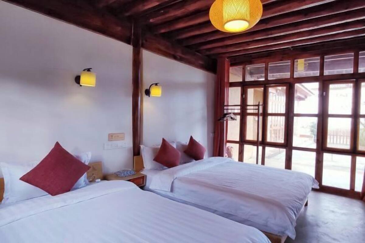 A hotel room with two beds, wooden beams, a large window, and wall-mounted lamps.
