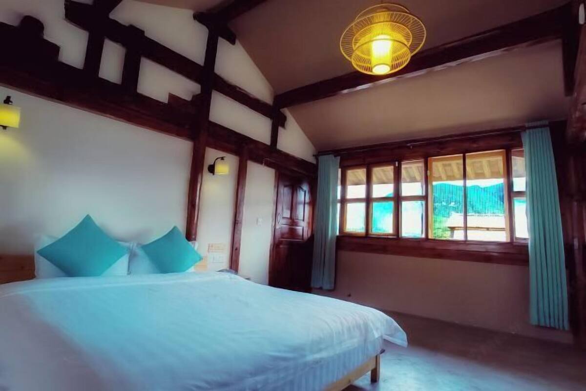 A bedroom with a bed, wooden beams, a window with curtains, and a hanging light fixture.