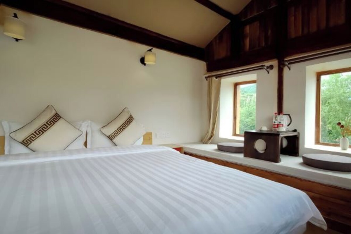 A hotel room with a bed, two pillows, a nightstand, a window with curtains, and a view of greenery outside.