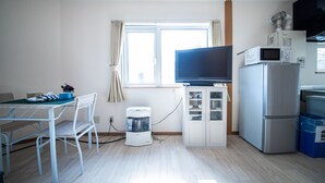 A small, well-lit room with a dining table, chairs, a television, a refrigerator, a microwave, and a window with curtains. - SUMIYOSHI HOUSE ROOMA (Otaru)