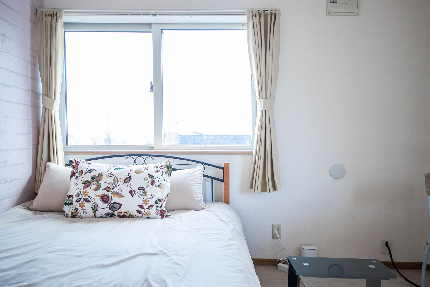 A bedroom with a bed, a window, curtains, a bedside table, and a power outlet.