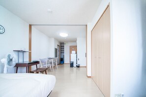 Et hotelværelse med seng, skrivebord, stol, ventilator og mikroovn i tekøkkenet.