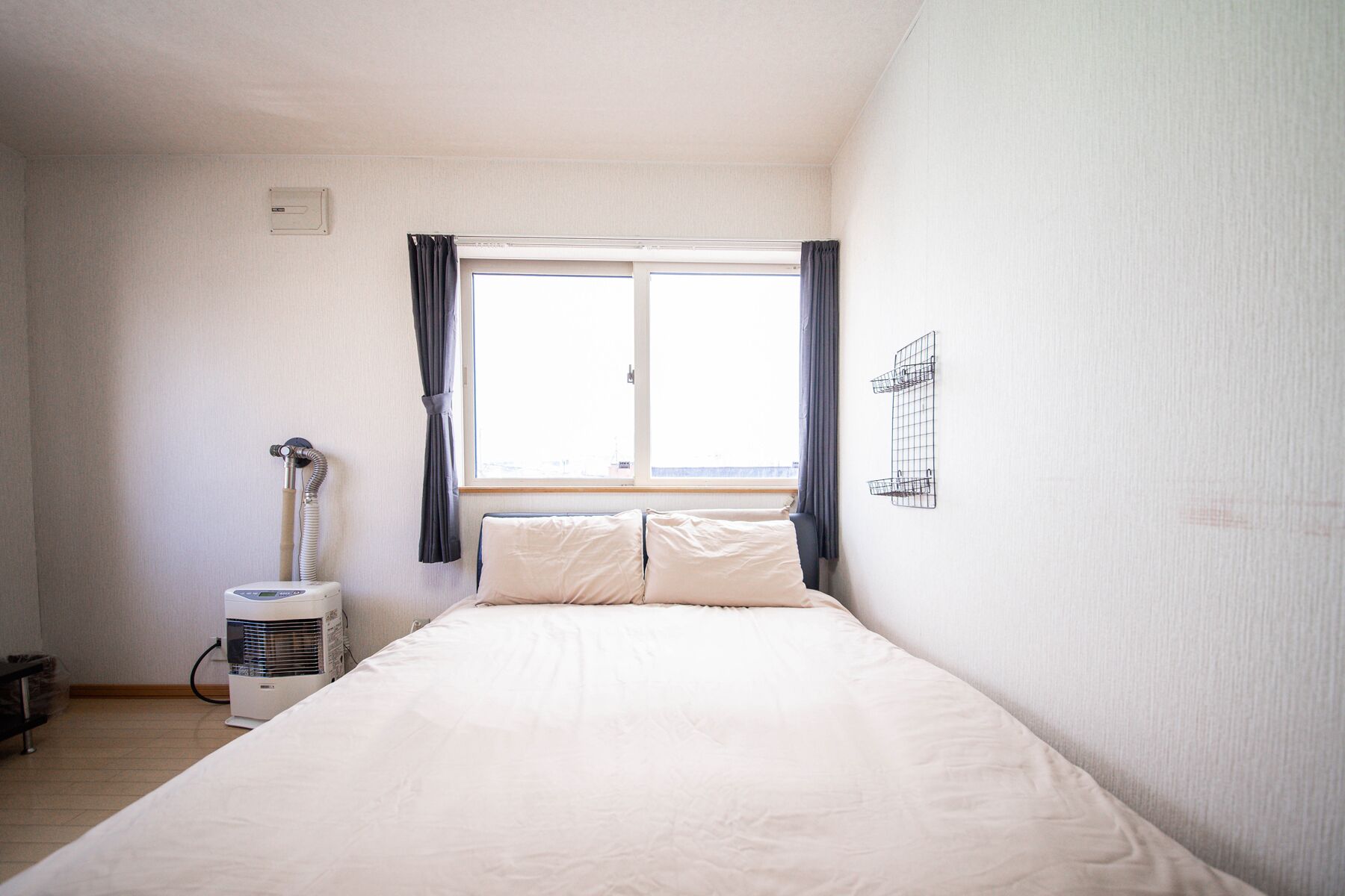 A small, well-lit bedroom with a single bed, a window with curtains, a radiator, and a wall-mounted shelf.