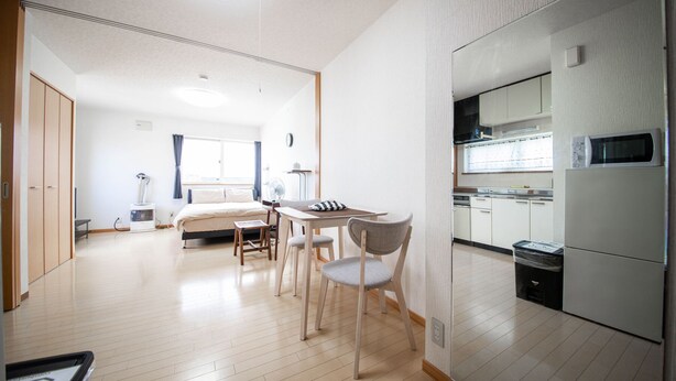 A modern hotel room with a bed, a dining area with a table and chairs, a kitchen with a refrigerator and microwave, and a large window with curtains. - SUMIYOSHI HOUSE ROOMB (Otaru)
