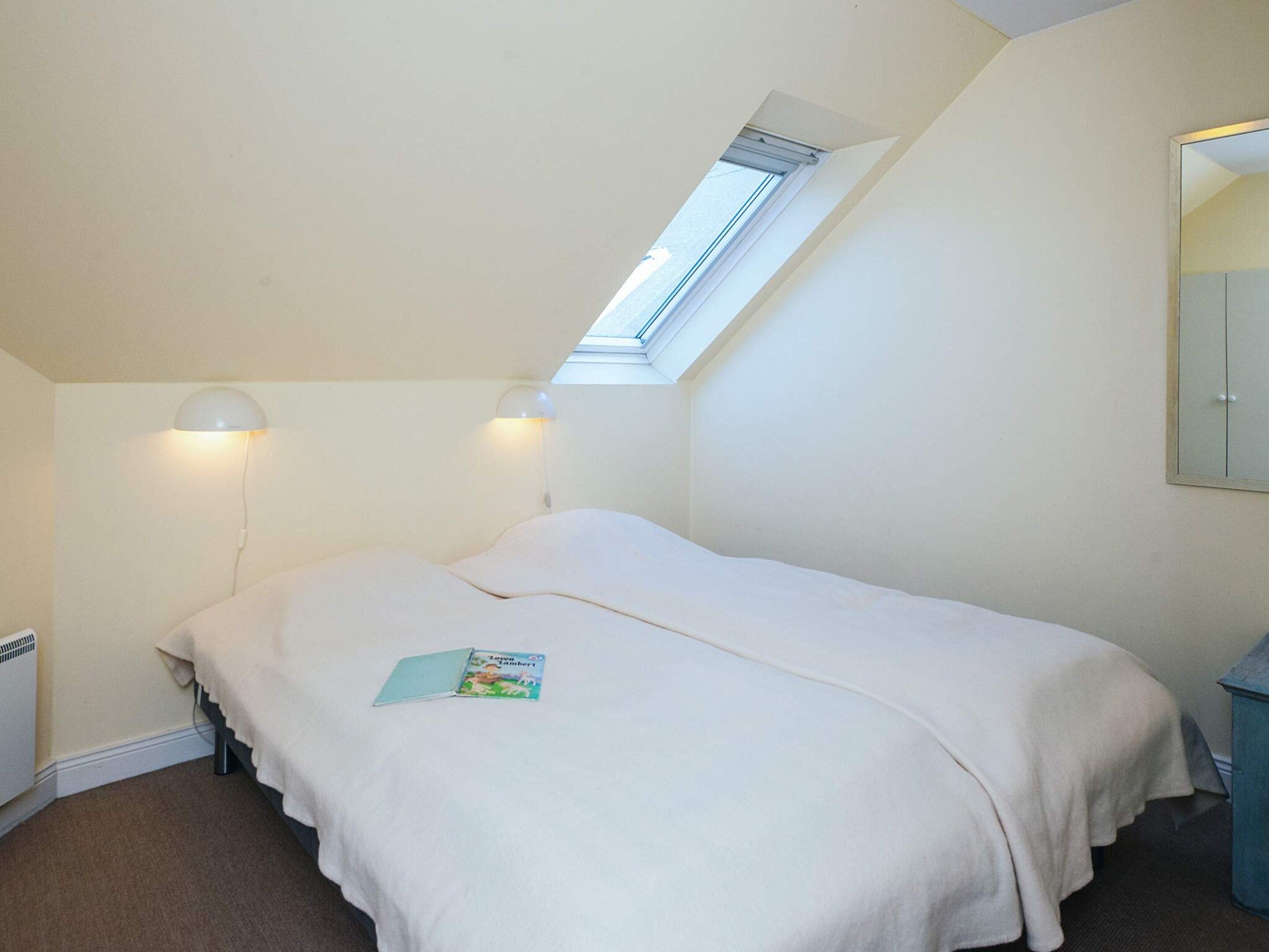 Ein Zimmer mit einem Einzelbett, einem Oberlicht, zwei Nachttischlampen, einem Nachttisch und einer kleinen Ablage.