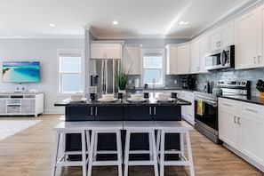 A modern kitchen with white cabinets, a central island with bar stools, stainless steel appliances, and a large window overlooking the ocean. - Spectacular home with Pool, New 8 seater Golf Cart, elevator, with easy access to beach (Oak Island)