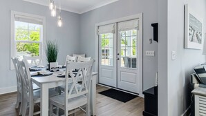 Ein Essbereich mit einem weißen Tisch und Stühlen, einer Zimmerpflanze und Blick ins Freie durch große Fenster.