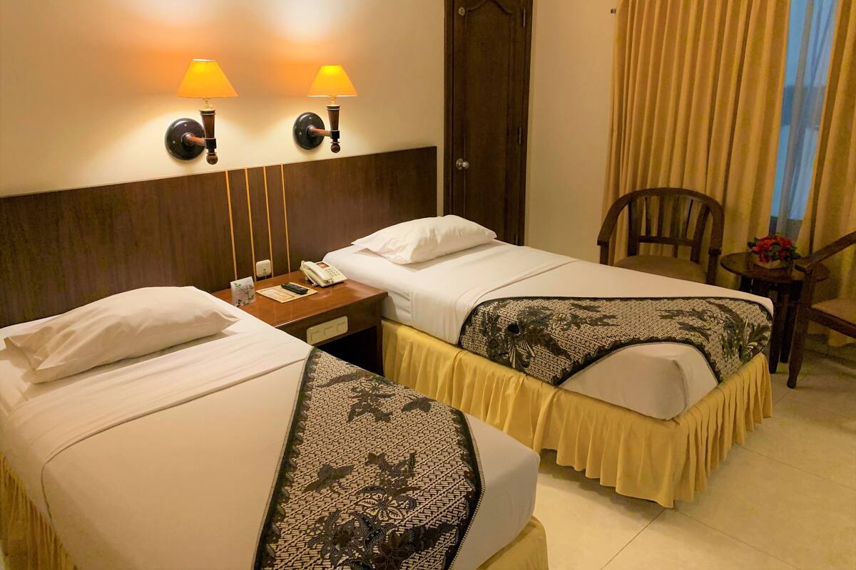 Een hotelkamer met twee bedden, een nachtkastje met telefoon, een stoel en een raam met gordijnen.