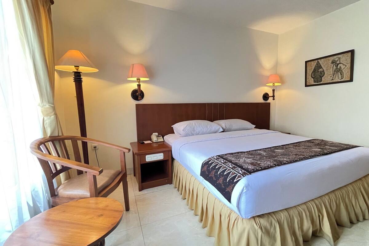 Een hotelkamer met een bed, nachtkastje, lamp, stoel en een schilderij aan de muur.