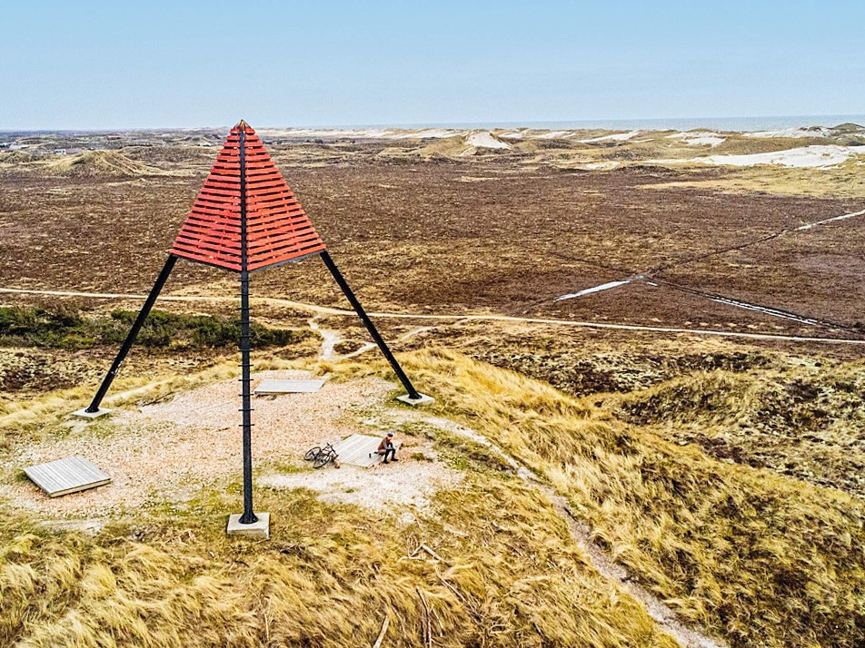 Une structure triangulaire rouge, dotée d’une base, se dresse dans un paysage aride, où la végétation est rare et le ciel est dégagé.