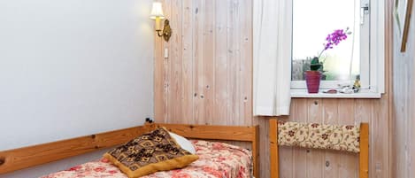 A small room with a bed, a chair, a window with a curtain, and a wall-mounted lamp.