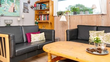 A living room with a wooden table, two black leather sofas, a bookshelf, and a window with plants.