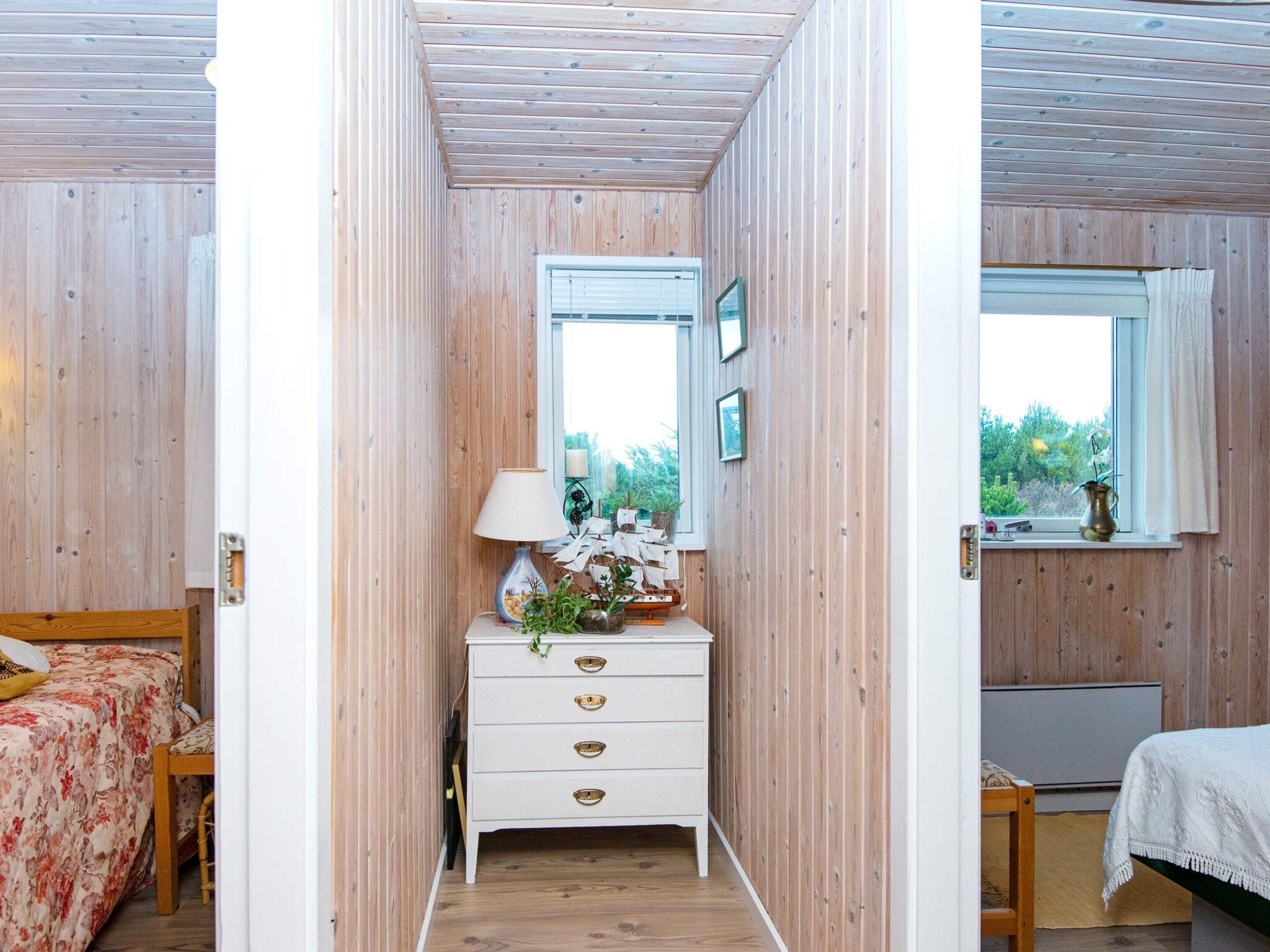 Et værelse med trævægge, en hvid kommode med skuffer, en seng med blomstret dynebetræk, et vindue med udsigt til grønne omgivelser og et lille bord med en lampe.