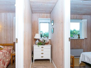 A room with wooden walls, a white dresser with drawers, a bed with a floral bedspread, a window with a view of greenery, and a small table with a lamp.