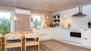 A modern kitchen with white cabinets, a wooden dining table with floral chairs, and a large window with curtains. - 7 Person Holiday Home in Hvide Sande-by Traum (Hvide Sande)