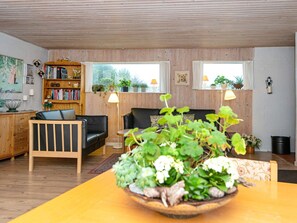 A cozy living room with a wooden floor, a sofa, a coffee table with a plant, a bookshelf, and two windows with curtains.